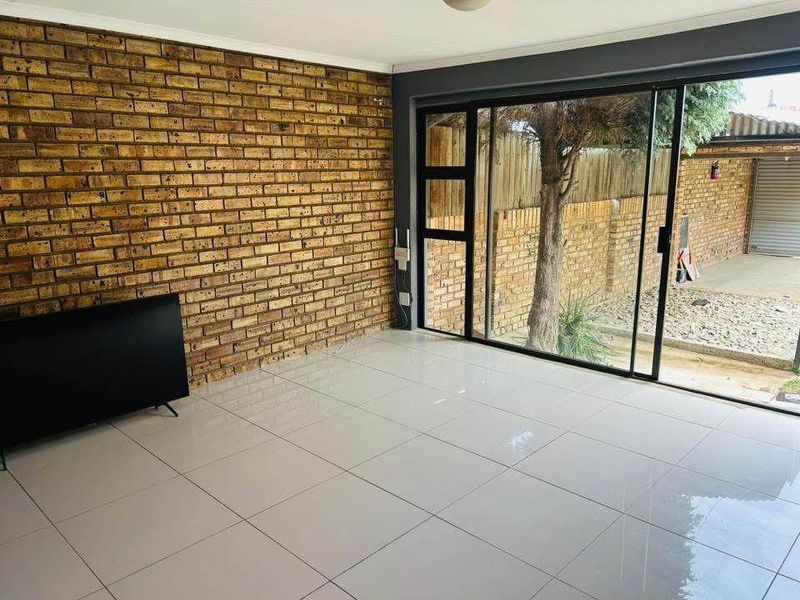 A bright, empty living room featuring a prominent exposed brick feature wall, large sliding glass doors, and modern light-colored floor tiles.