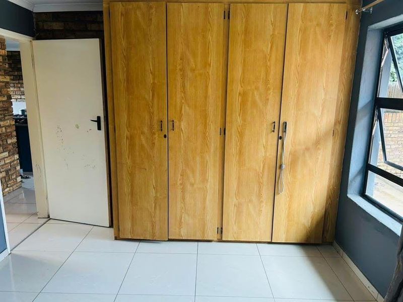A bedroom featuring tiled flooring, an aluminium window, and built-in wooden cupboards, offering a functional space ready for personal touches.