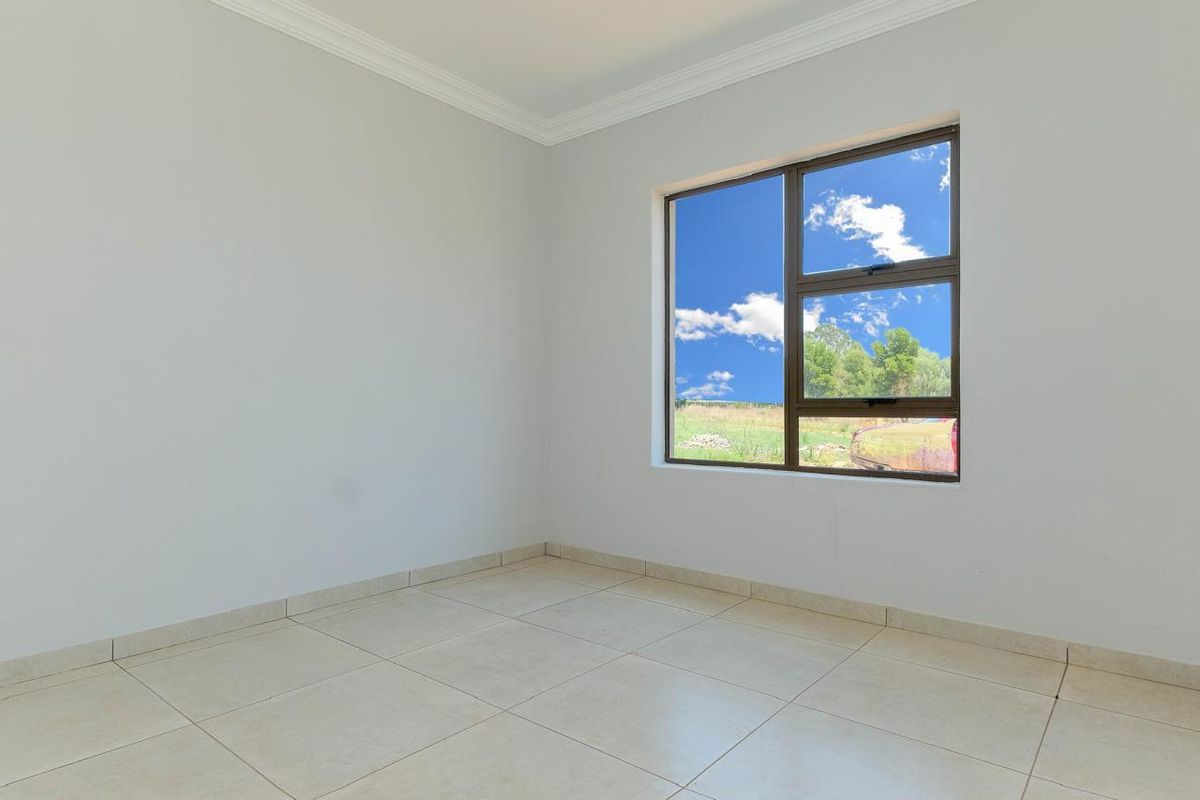 A bright, empty bedroom featuring neutral tiled flooring, clean white walls, and an aluminium-framed window with a view of the surrounding landscape.