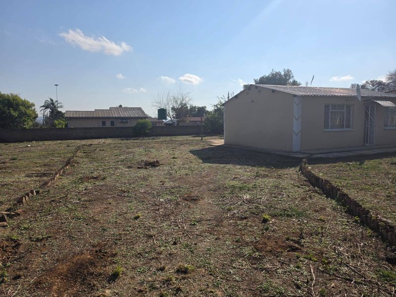 A view of the rear yard featuring a large open space, brick boundary wall, and the side elevation of the house with security features.