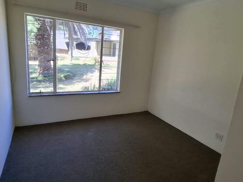 An empty bedroom featuring a large steel-framed window, carpeted flooring, and neutral walls, offering a blank canvas for renovation or staging.