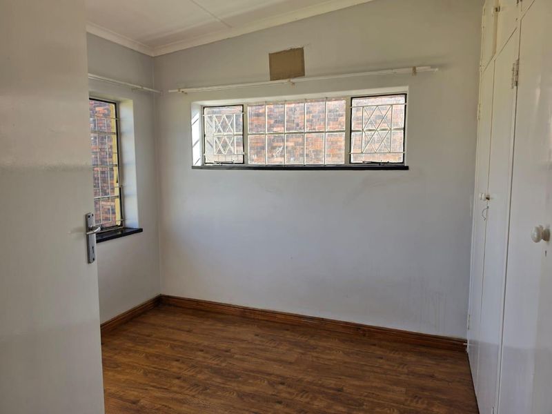 An empty bedroom featuring laminate flooring, built-in cupboards, and windows fitted with security bars, requiring minor cosmetic updates to the walls.