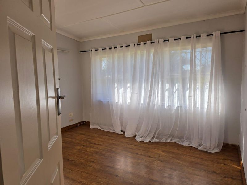 A bright, empty bedroom featuring laminate flooring, neutral walls, and a large window fitted with a curtain rod and security gate.