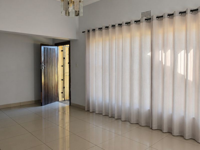 A bright and spacious living area featuring neutral tiled flooring, large windows with curtain tracks, and a secure entrance door.