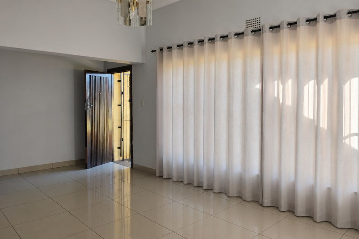 A bright and spacious living area featuring neutral tiled flooring, large windows with curtain tracks, and a secure entrance door.