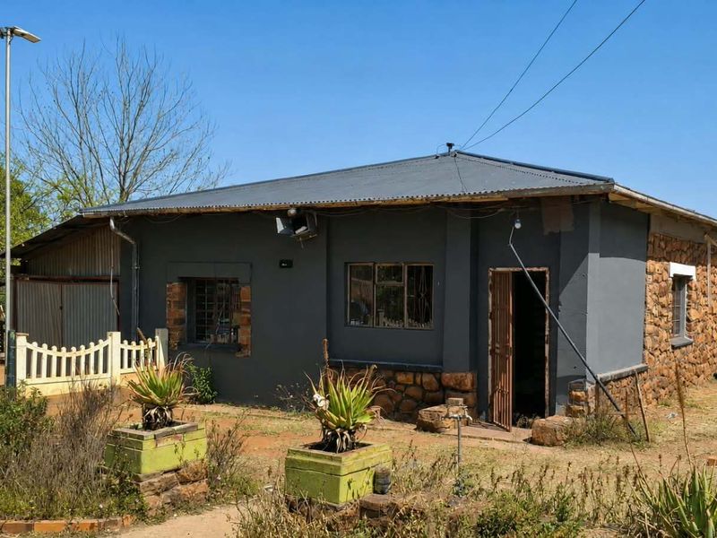 A single-story house featuring a combination of stone masonry and painted walls with a corrugated metal roof and security gate entrance.