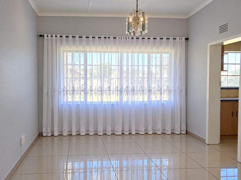 A bright and spacious living area featuring polished tiled floors, a decorative crystal chandelier, and a large window with security bars.
