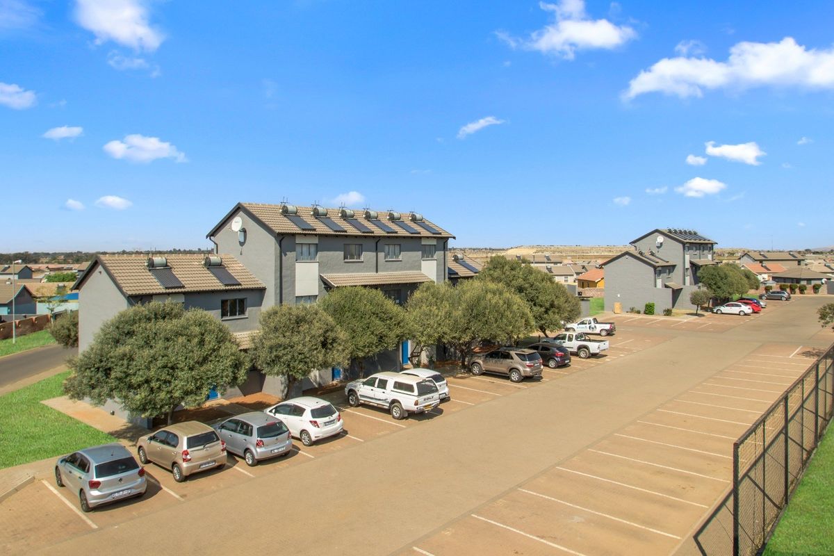 Aerial view of a modern apartment complex featuring grey facades, tiled roofs with solar water heaters, and an extensive paved parking area.