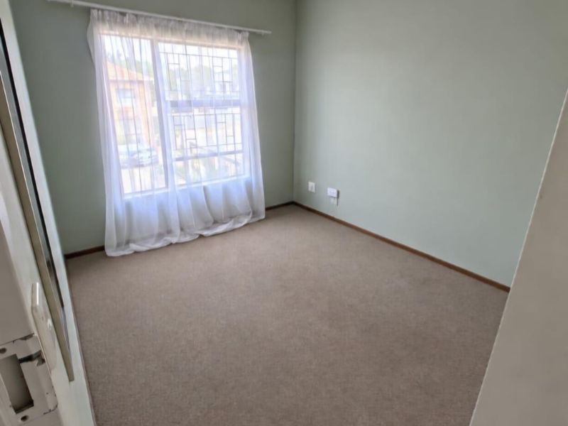 A clean, empty bedroom featuring neutral green walls, carpeted flooring, and a window fitted with security bars.