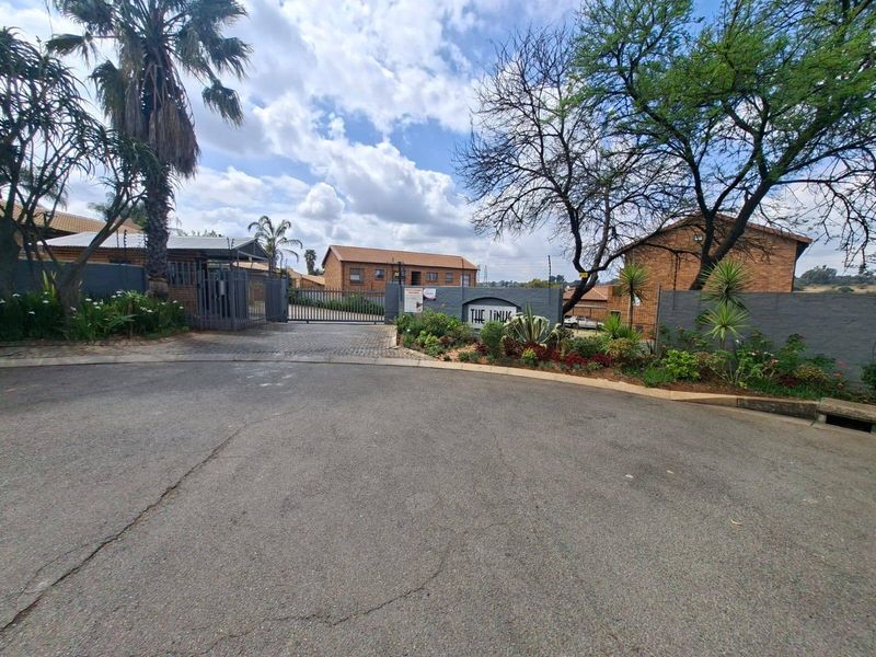Front entrance view of the apartment complex featuring a secure gated access point, perimeter wall with electric fencing, and established garden beds.