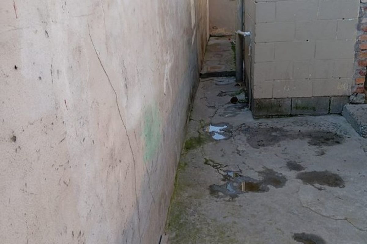 A narrow side alleyway featuring concrete paving and masonry walls, showing signs of wear, cracking, and poor drainage.