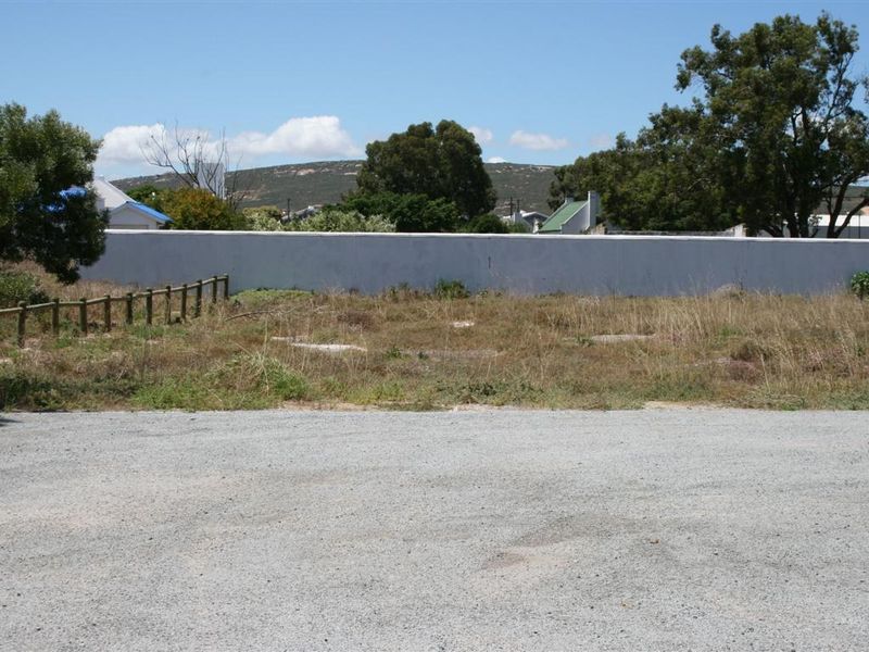 A vacant residential plot featuring a gravel driveway, a perimeter boundary wall, and established trees in the background.
