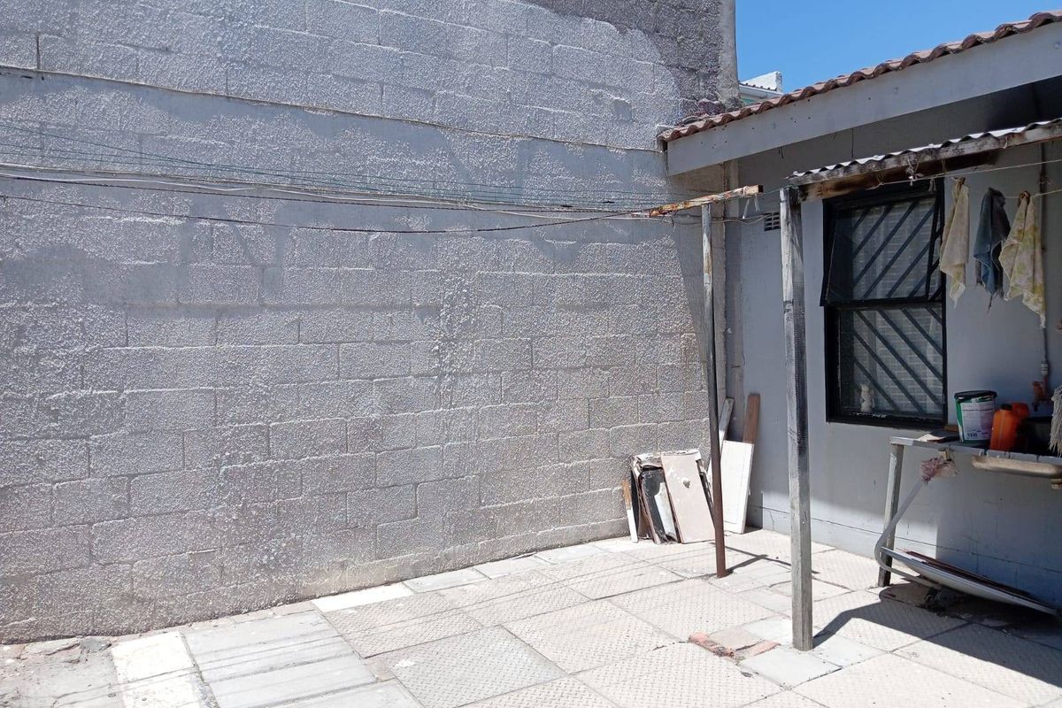 A rear exterior utility area featuring a paved courtyard, a large masonry wall, and a basic metal-framed structure with a sink.