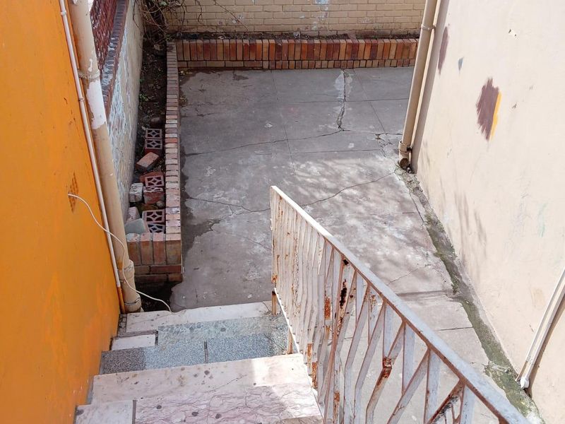 An outdoor rear area featuring concrete paving, a brick planter box, and a staircase with a rusted metal railing.