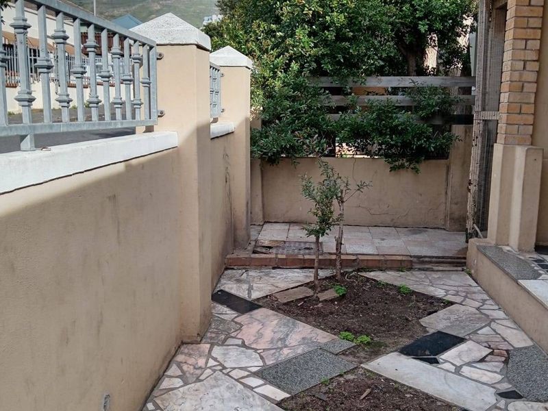 A paved courtyard area featuring a mix of stone tiles and small garden beds, enclosed by a boundary wall with metal railings.