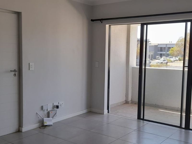 A bright, empty living area featuring neutral tiled flooring and a large sliding glass door providing access to an outdoor balcony.