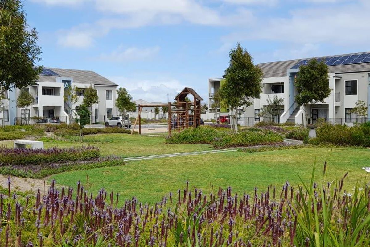 A well-maintained communal garden area between apartment blocks, featuring landscaped lawns, flower beds, a paved pathway, and a children's playground.