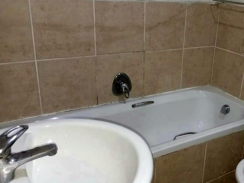 A compact bathroom featuring a bathtub, pedestal basin, and neutral ceramic wall tiling, currently showing signs of wear and requiring cosmetic updates.
