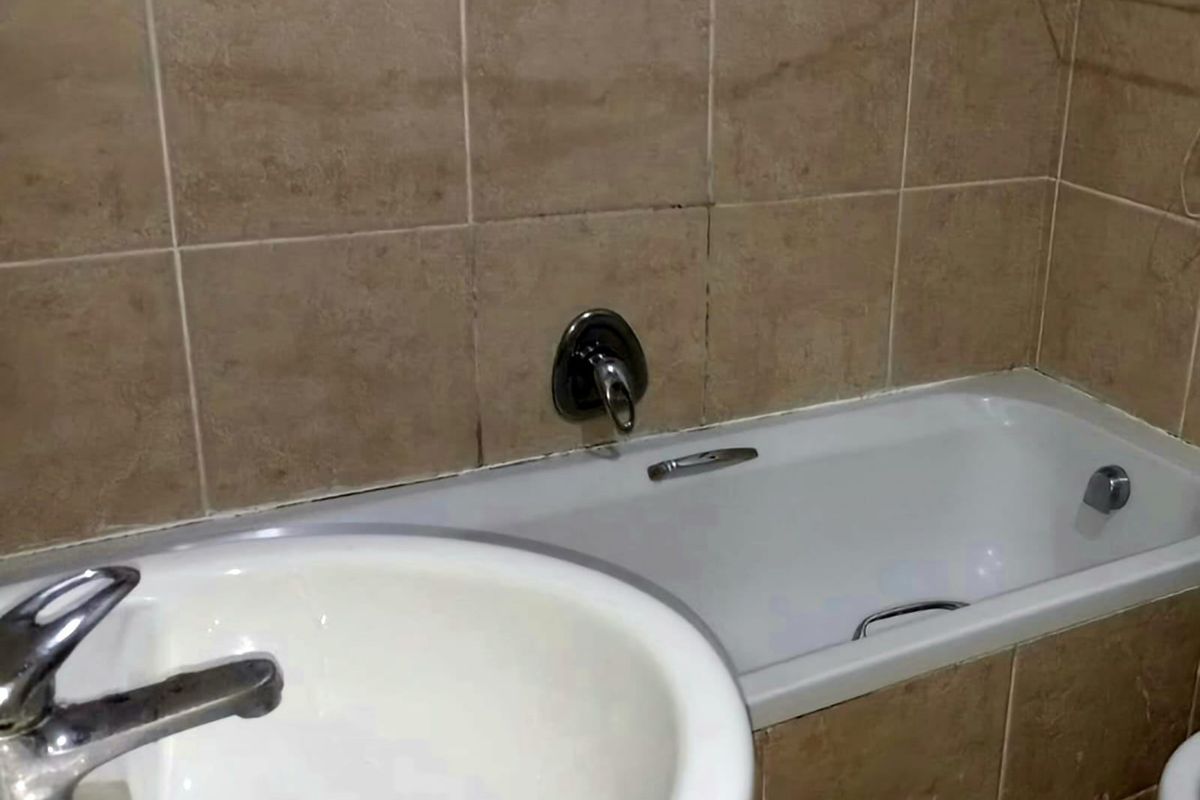 A compact bathroom featuring a bathtub, pedestal basin, and neutral ceramic wall tiling, currently showing signs of wear and requiring cosmetic updates.