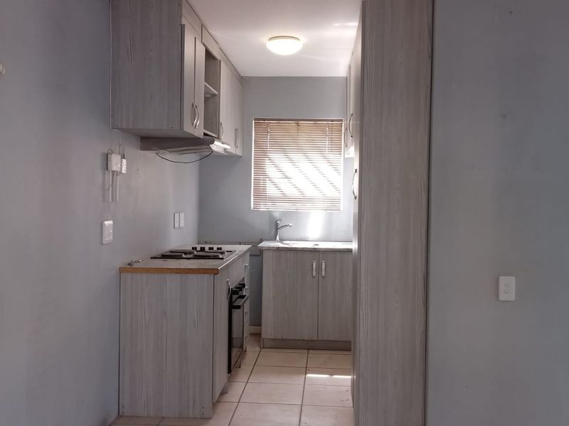 A compact, functional kitchen featuring light wood-grain cabinetry, built-in oven and hob, tiled flooring, and a window with venetian blinds.