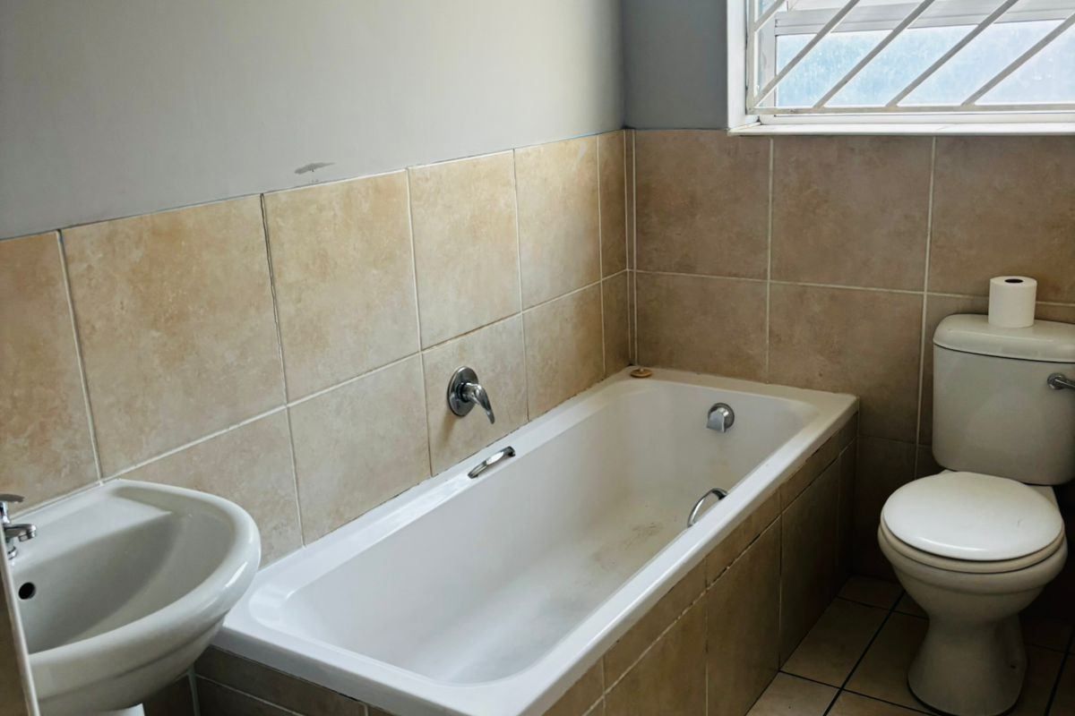 A functional bathroom featuring a pedestal basin, built-in bathtub, and toilet with neutral wall tiling and a window with security bars.