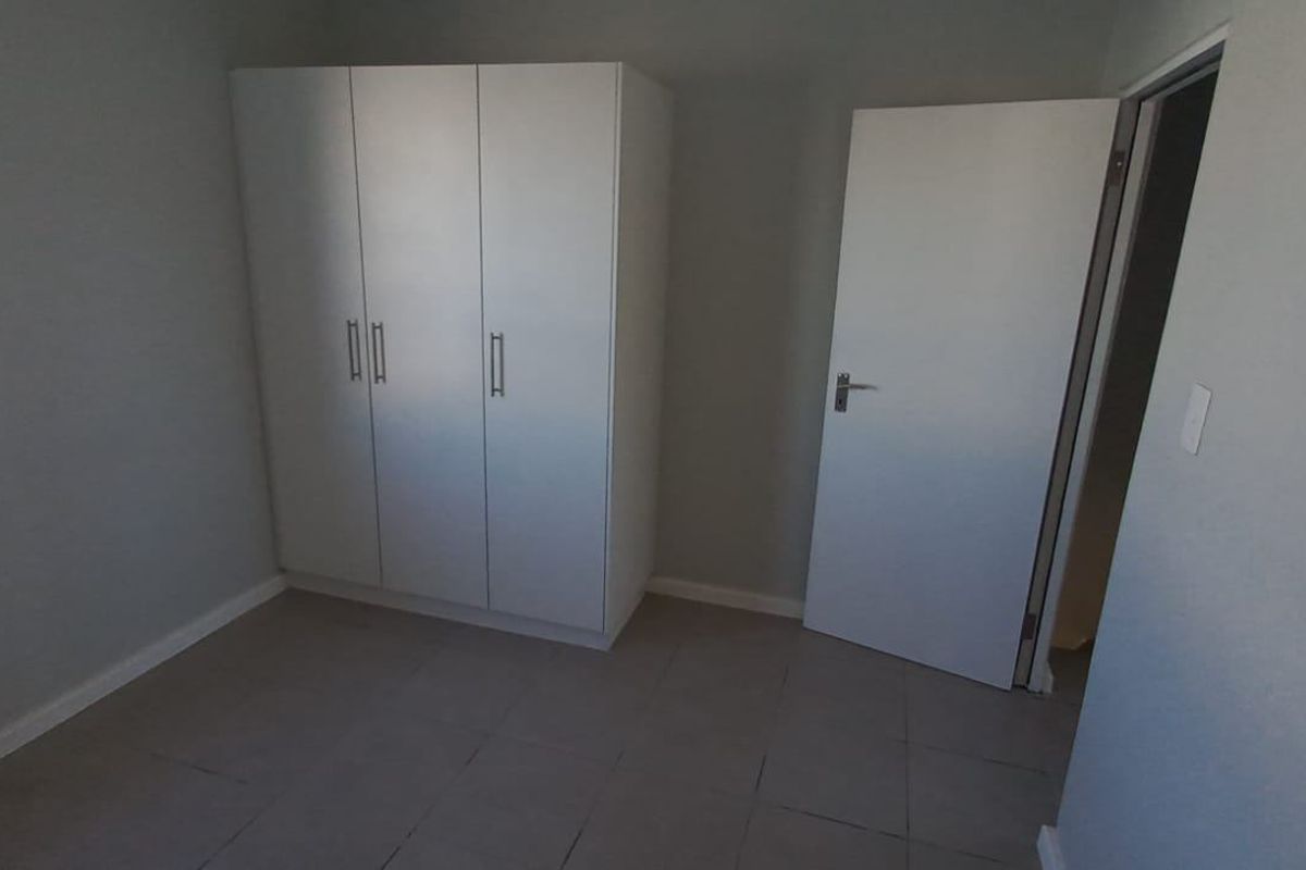 A clean, neutral bedroom featuring tiled flooring, a built-in wardrobe, and a window providing natural light.