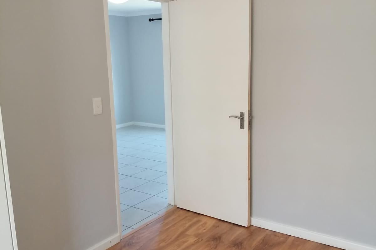 A clean, empty bedroom featuring light grey walls, laminate wood-look flooring, and a white internal door leading to an adjacent tiled room.