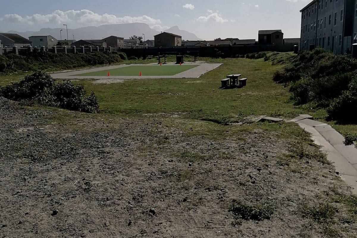 An exterior view of the apartment complex grounds featuring a paved walkway, perimeter wall, and a communal recreational area with unmaintained landscaping.