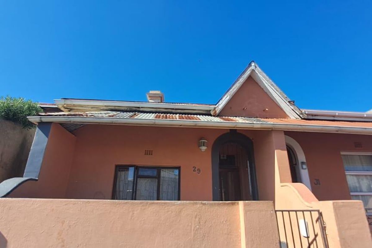 Front elevation of a residential property featuring a corrugated iron roof, brick boundary wall, and gated entrance, showing signs of significant exterior wear.