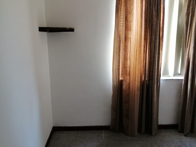 A simple, empty bedroom featuring tiled flooring, a wall-mounted shelf, and window curtain rails.