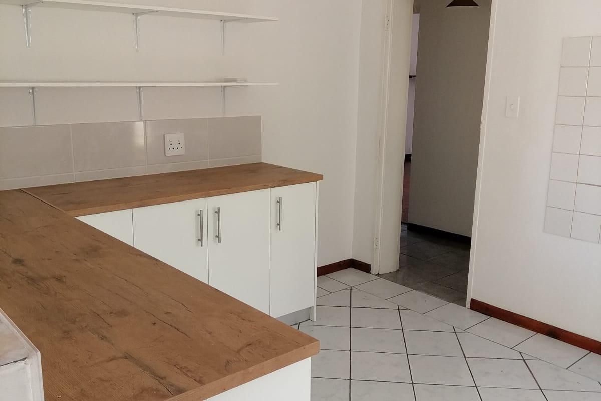 A clean, empty kitchen area featuring modern white cabinetry, wooden countertops, tiled flooring, and wall-mounted shelving, offering a functional and neutral space.