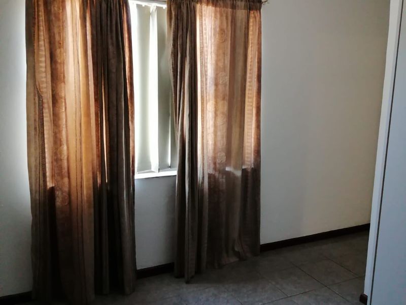 A clean, empty bedroom featuring neutral tiled flooring, a built-in cupboard, and natural light through a window with curtains.