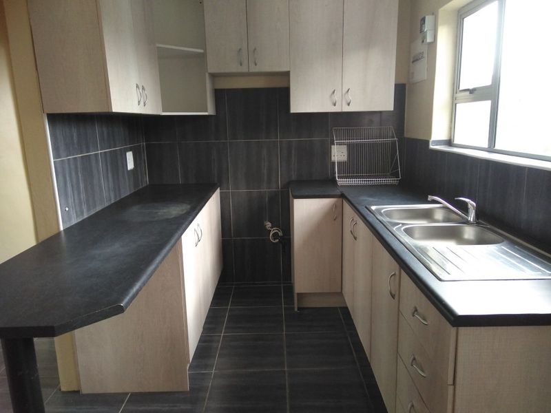 A functional kitchen featuring light-toned cabinetry, dark laminate countertops, a double stainless steel sink, and dark tiled splashbacks and flooring.