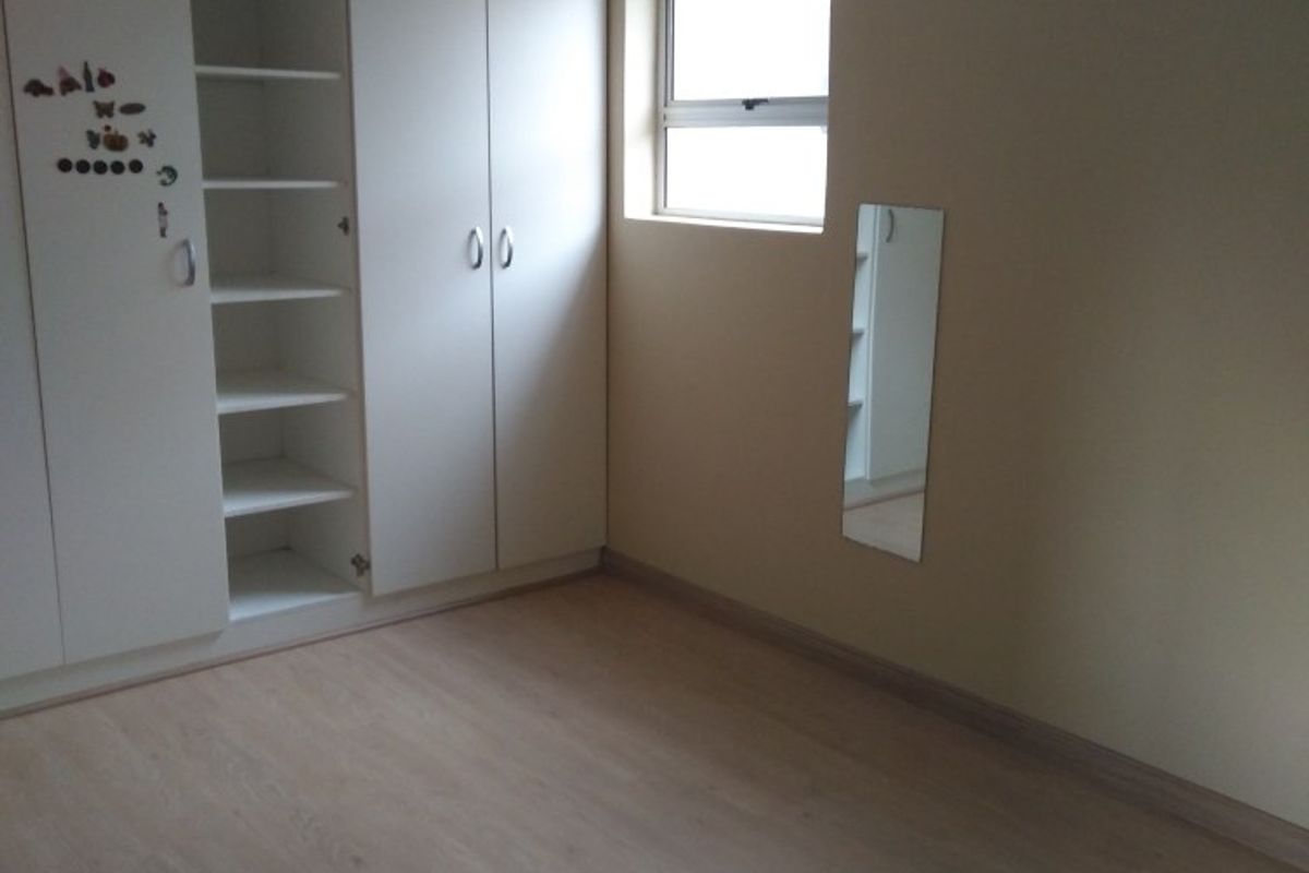 A clean, empty bedroom featuring light-toned laminate flooring, built-in white cupboards, and a window with a curtain rod.