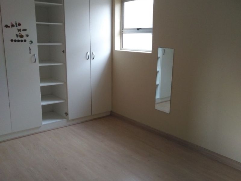 A clean, empty bedroom featuring light-toned laminate flooring, built-in white cupboards, and a window with a curtain rod.