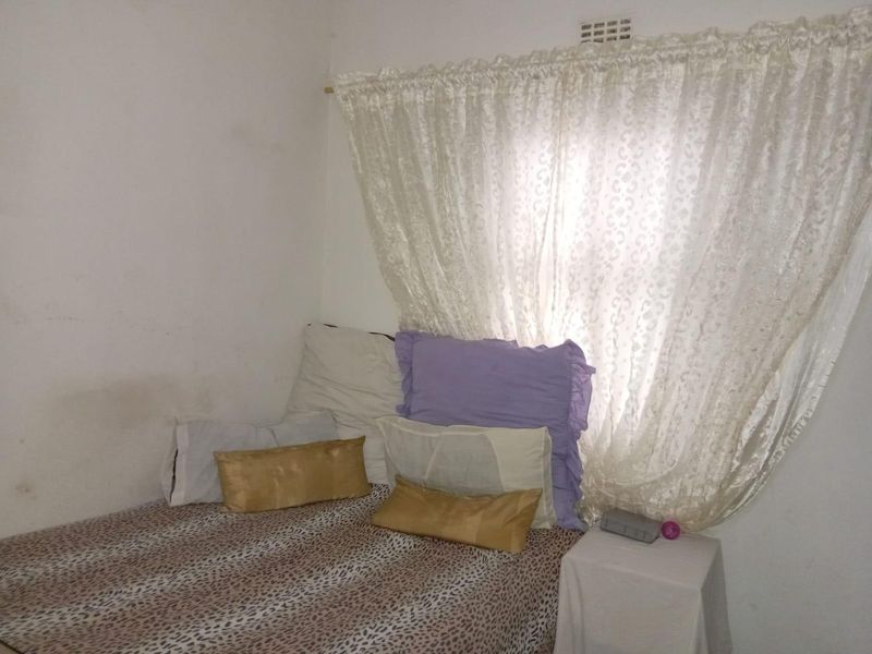 A compact bedroom featuring a window with curtains and a wall-mounted air vent, requiring cosmetic updates to the wall finishes.