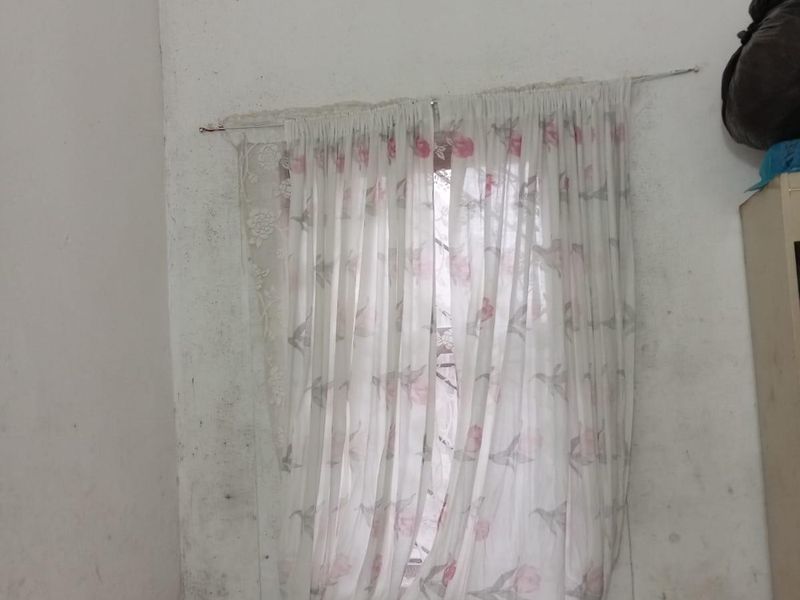 A bedroom corner featuring a window with floral curtains, showing significant wall surface wear and staining requiring maintenance.