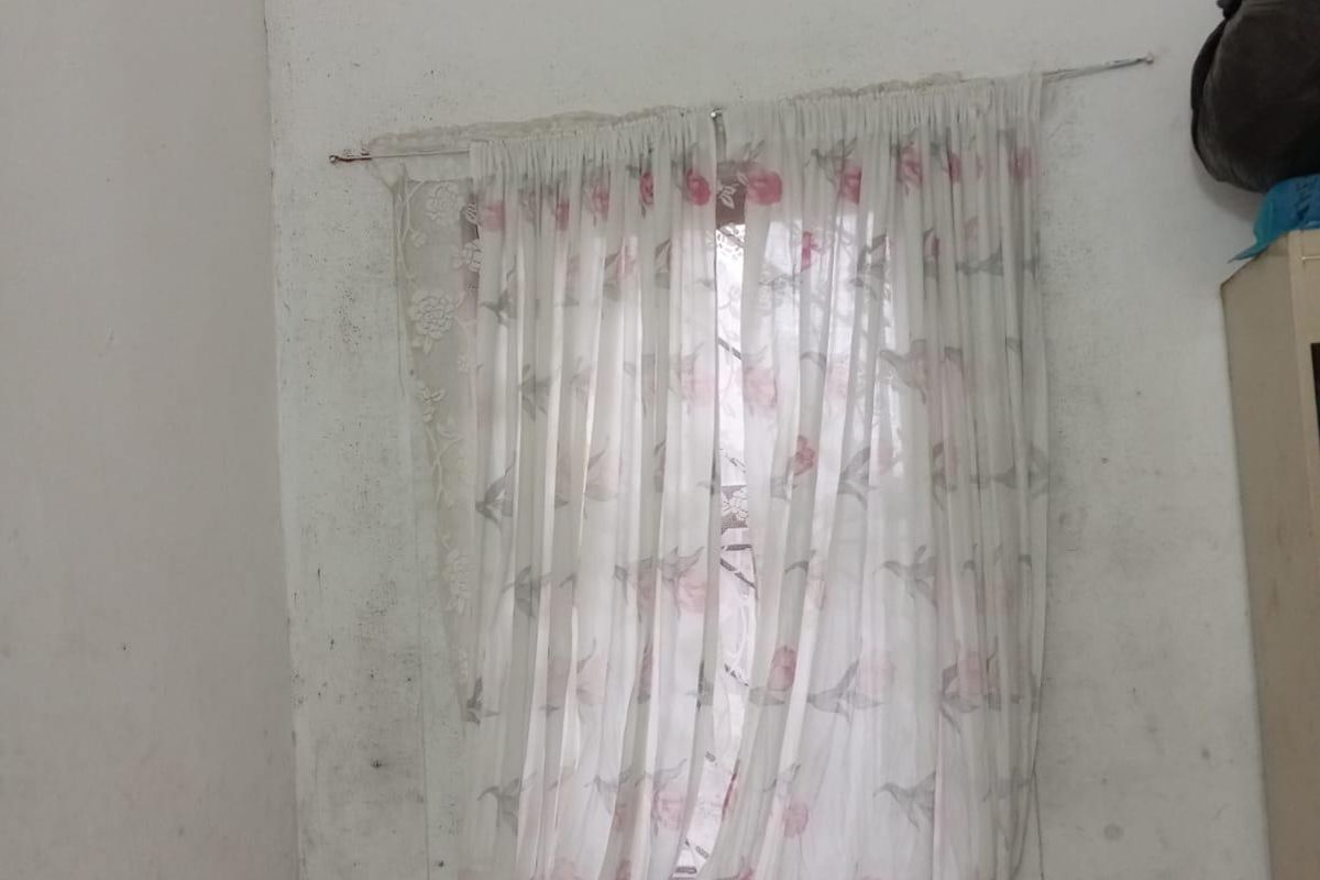 A bedroom corner featuring a window with floral curtains, showing significant wall surface wear and staining requiring maintenance.