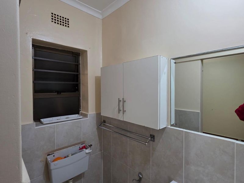 A functional bathroom featuring a wall-mounted toilet, ceramic basin, vanity storage cabinet, and tiled wall finishes with a louvered window for ventilation.