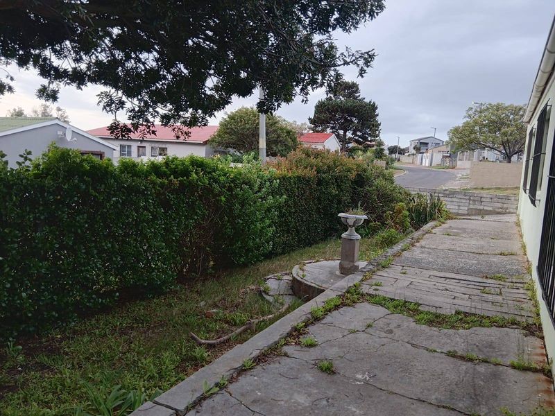 A rear exterior view showing a concrete pathway, retaining wall, and established garden hedging in a residential setting.