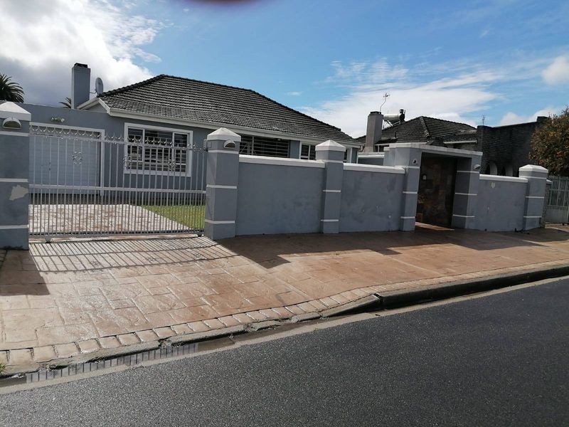 Front exterior view of a single-story house featuring a perimeter wall, metal gate, paved driveway, and tiled roof with solar water heating system.