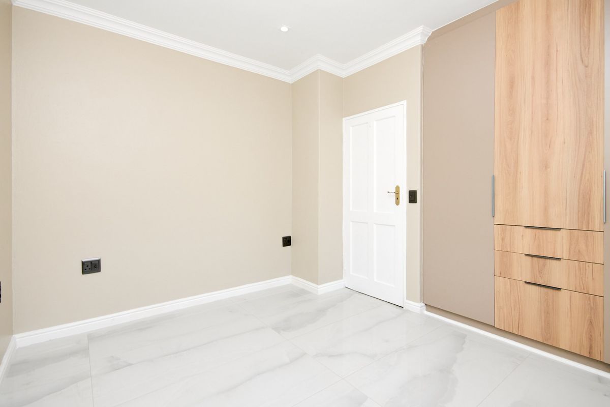 A bright, modern bedroom featuring neutral walls, high-quality tiled flooring, crown molding, and extensive built-in cabinetry with a mix of wood and matte finishes.