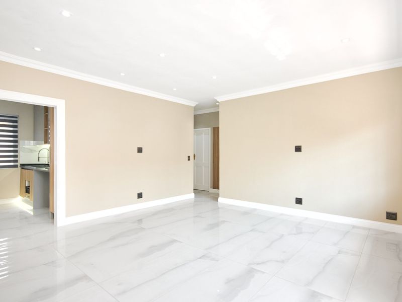 A bright, empty living room featuring polished marble-look porcelain floor tiles, recessed downlights, and neutral wall finishes, with a view into the adjacent kitchen.
