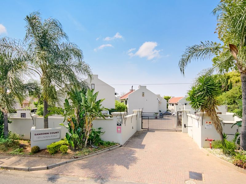 Front entrance view of Sandown Court townhouse complex featuring a paved driveway, secure metal gate, and perimeter walling.