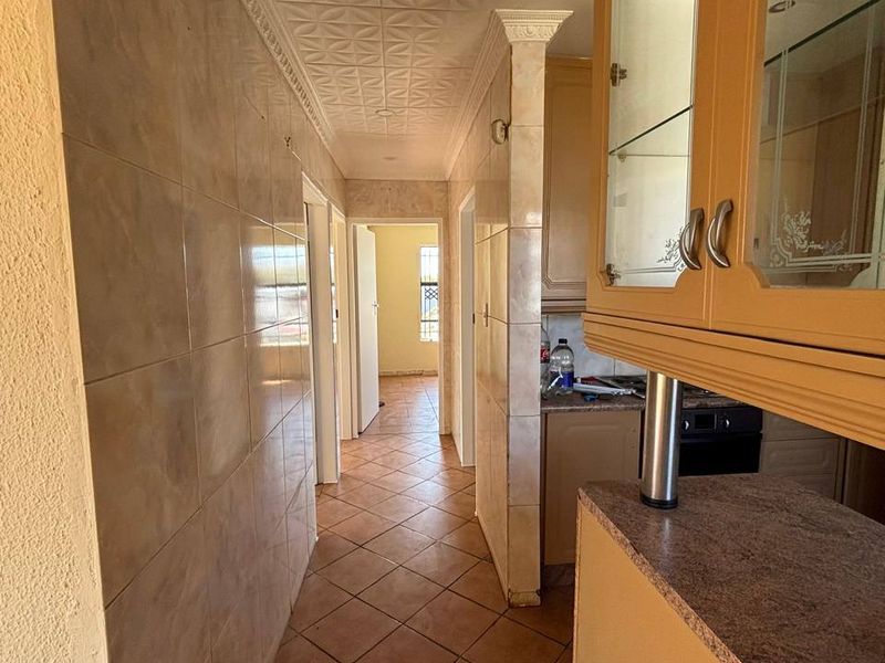 A dated kitchen area featuring tiled walls, ceramic floor tiles, and traditional cabinetry with glass-fronted display units, leading into a hallway.