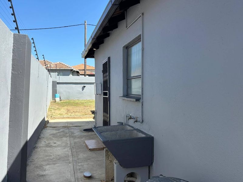 Rear exterior view showing a paved walkway, perimeter wall with electric fencing, and a fixed concrete laundry trough attached to the building exterior.