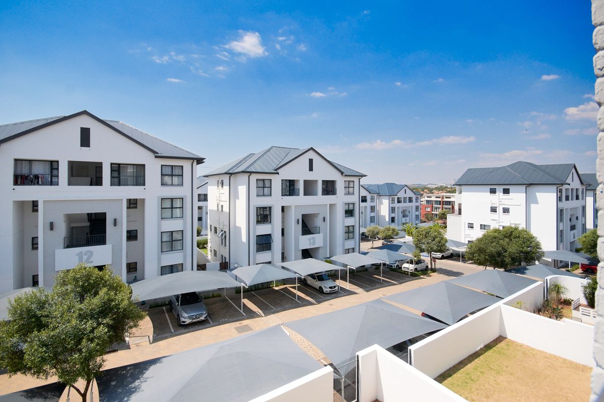 An elevated aerial view of a modern apartment complex featuring multiple residential buildings, paved driveways, and designated covered parking bays.