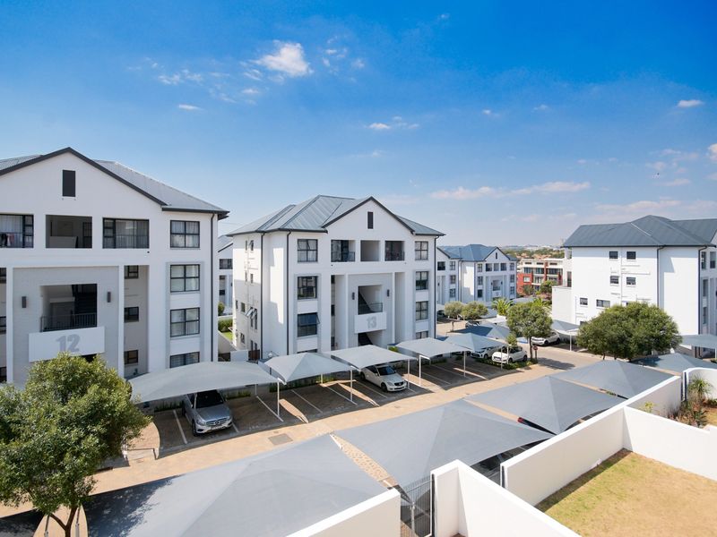 An elevated aerial view of a modern apartment complex featuring multiple residential buildings, paved driveways, and designated covered parking bays.