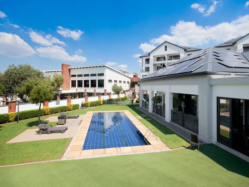 A well-maintained communal pool area featuring a tiled swimming pool, paved patio space, and landscaped garden surroundings within an apartment complex.
