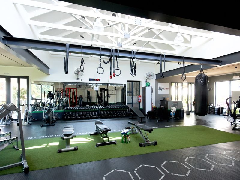 A spacious, well-equipped gym featuring exposed roof trusses, rubber flooring, artificial turf sections, and a large mirrored wall for training.
